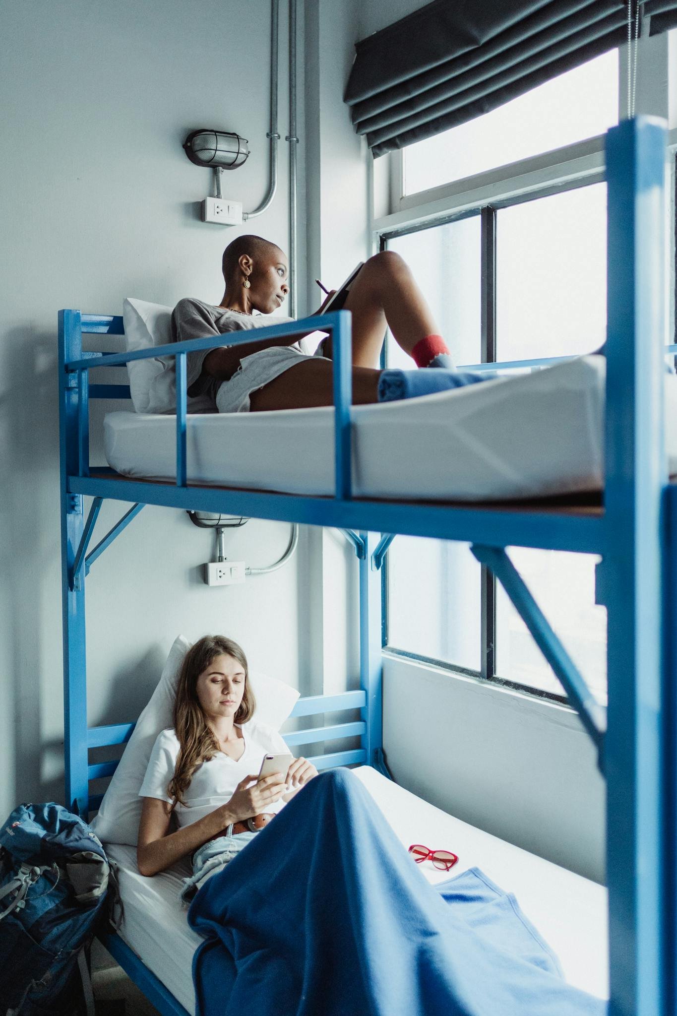 Two women reclining in a bright hostel room, enjoying leisure time on bunk beds.