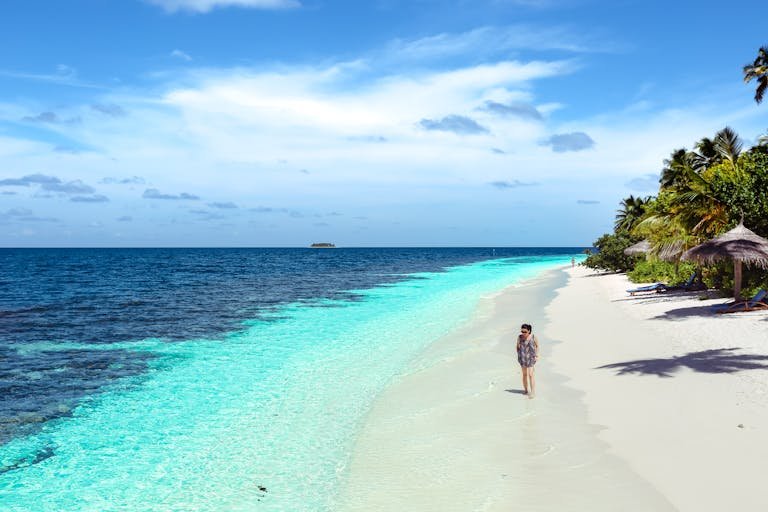 Solo Travel Koh Tao Guide. A woman strolling along a pristine tropical beach during the day, surrounded by clear blue waters and lush greenery.
